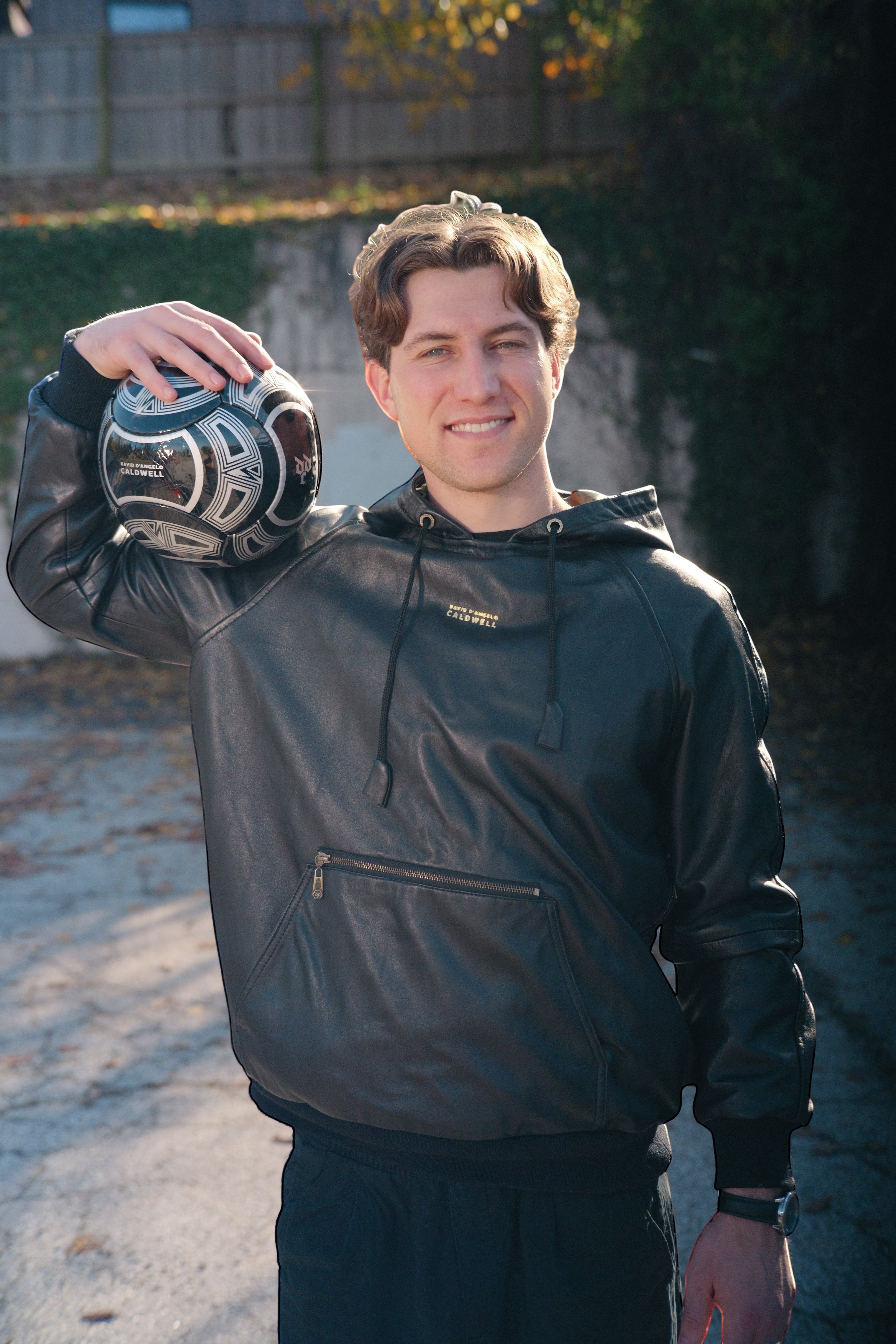 David Dangelo Caldwell black lamb leather reversible hoodie with oversized hood and zipper pockets, worn by a man holding a soccer ball.